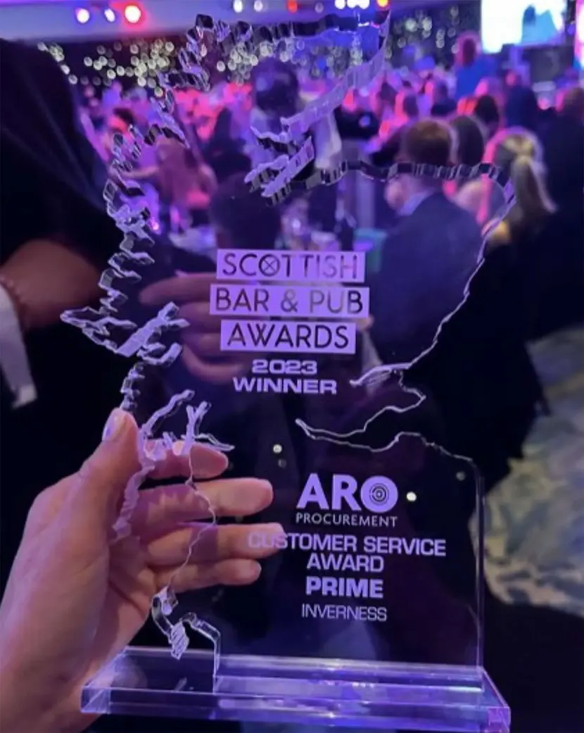 A hand holding an award shaped like Scotland, labeled "Scottish Bar & Pub Awards 2023 Winner" with "ARO Procurement Customer Service Award Prime Inverness" below. The crowded background showcases a seamlessly executed evening by top-notch Event Management professionals.
