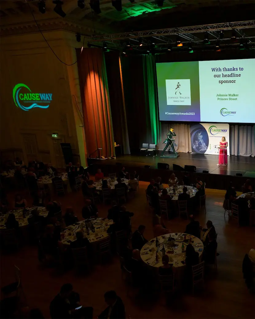 A speaker is on stage at the Causeway Awards event, with attendees seated at round tables. Sponsor logos are displayed on screens and a wall above the stage, showcasing expert event management.