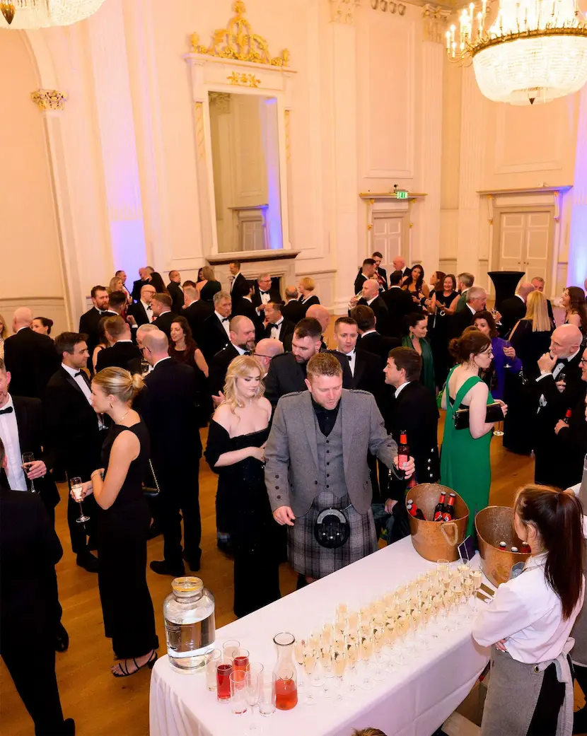 A large group of people in formal attire gathered in an elegant, chandelier-lit room. They are mingling and conversing, with a few standing near a table with glasses of beverages—an impeccable example of exceptional Event Planning.