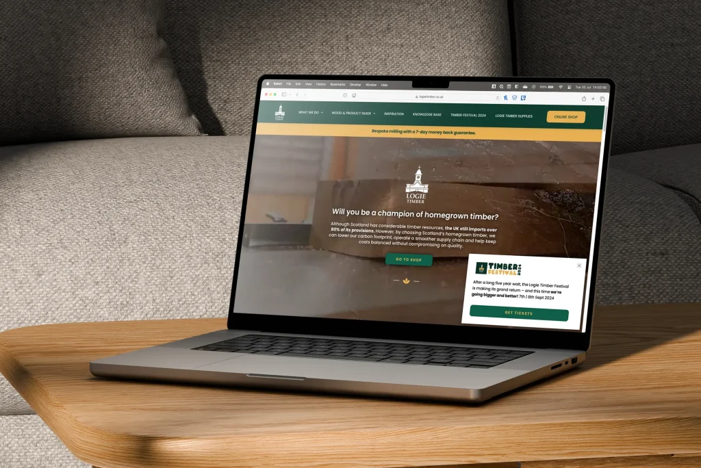 A laptop on a wooden table displays a bespoke website about homegrown timber champions. The screen shows text and images related to timber projects and an option to enroll.