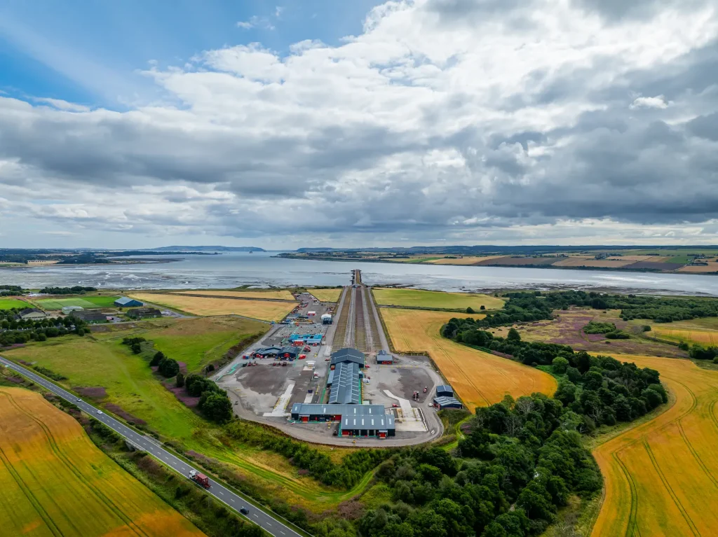 Aerial view of a rural landscape featuring a Green Freeport with a long, narrow structure extending towards a body of water, surrounded by fields and trees under a cloudy sky.