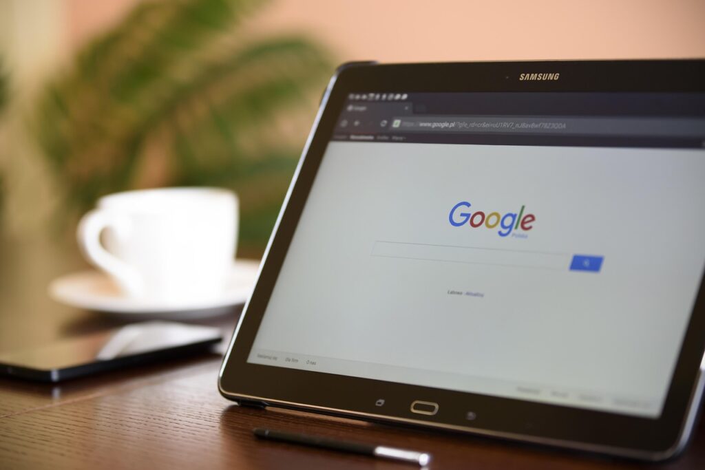 A tablet screen showing Google search engine's homepage with a white coffee cup, a smartphone and a white coffee cup in the background
