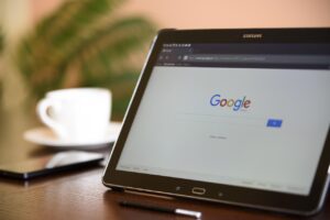 A tablet screen showing Google search engine's homepage with a white coffee cup, a smartphone and a white coffee cup in the background